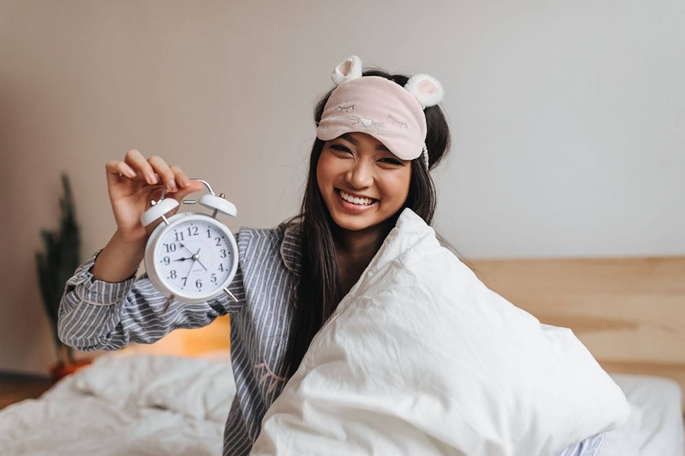 A woman with a comforter wrapped around her is holding an alarm clock