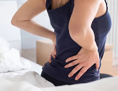 A person sitting on the bed with their hand on the back signifying pain