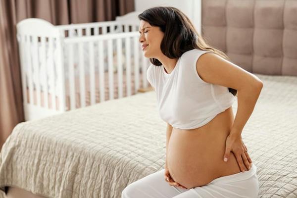 A pregnant woman sitting on the bed with one hand on her belly and the other on her lower back agonizing in pain