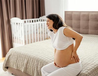 A pregnant woman sitting on the bed with one hand on her belly and the other on her lower back agonizing in pain