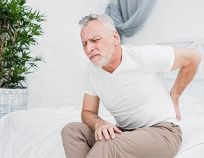 A man sitting bent forward on the bed with his hand on his back signifying lower back pain