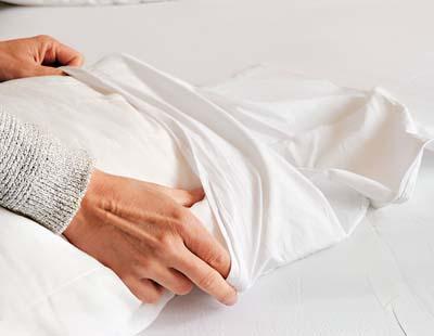 An individual covers a white pillow with a protective pillow cover on top of a well-arranged bed