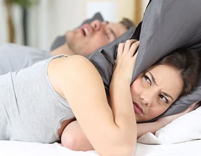 A woman covering her ears with pillows, unable to sleep due to the loud snoring noises of her partner
