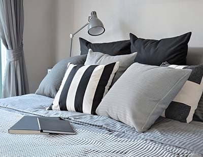 A couple of pillows neatly placed on a bed, with a lamp and notebooks kept on the side