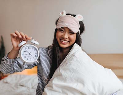 A woman with a comforter wrapped around her is holding an alarm clock