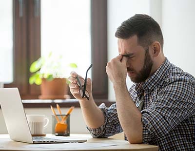 A man sits at a desk with a laptop, holding his glasses and rubbing his eyes, appearing stressed or fatigued