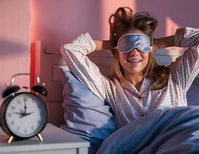 A woman with eye mask on wakes up on her bed, with an alarm clock on the night stand