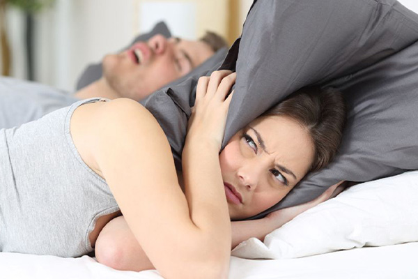 A woman covering her ears with pillows, unable to sleep due to the loud snoring noises of her partner