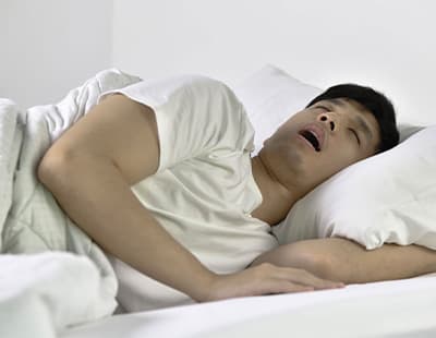 A man sleeping on his side with his mouth open and hand tucked under a pillow, indicating sleep apnea