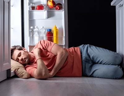 A man sleeping on the floor in the kitchen near an open refrigerator