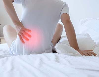 A man in pain with his arm on his lower back, sitting on the bed.