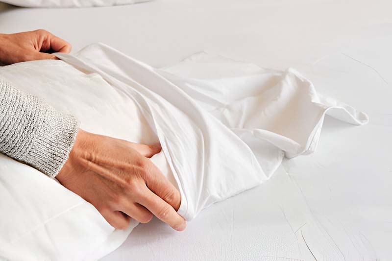 An individual covers a white pillow with a protective pillow cover on top of a well-arranged bed