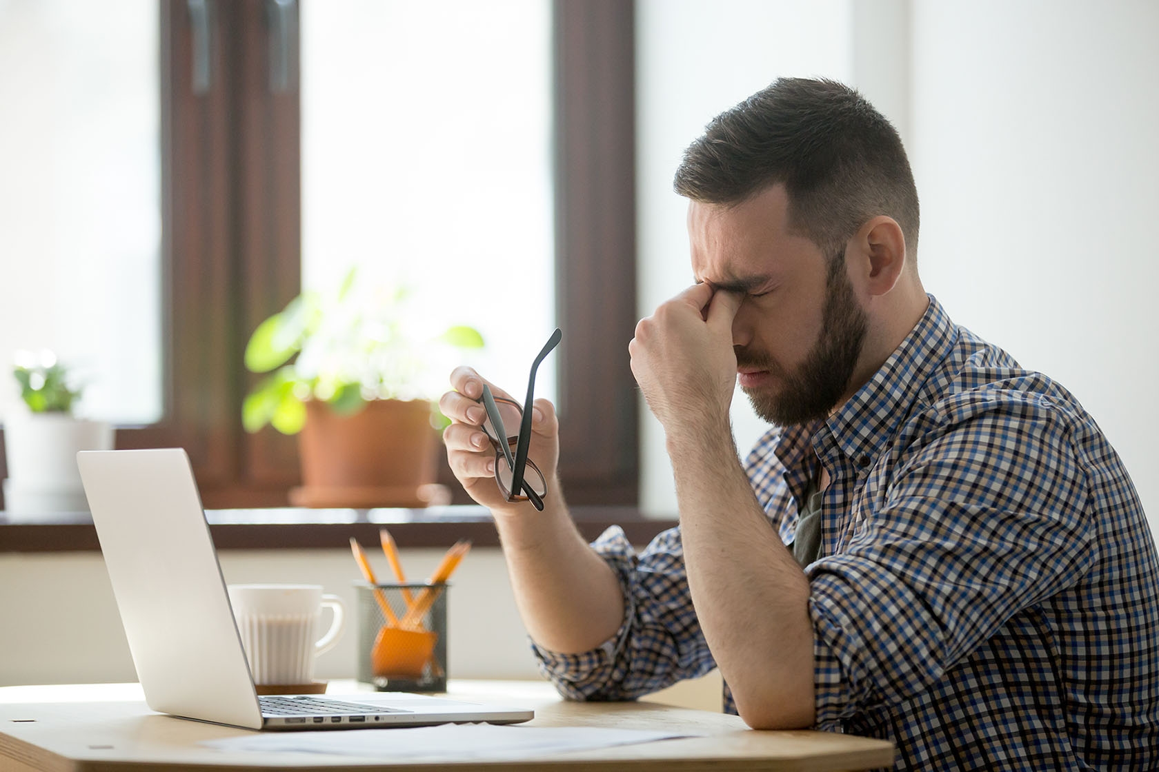 A man sits at a desk with a laptop, holding his glasses and rubbing his eyes, appearing stressed or fatigued