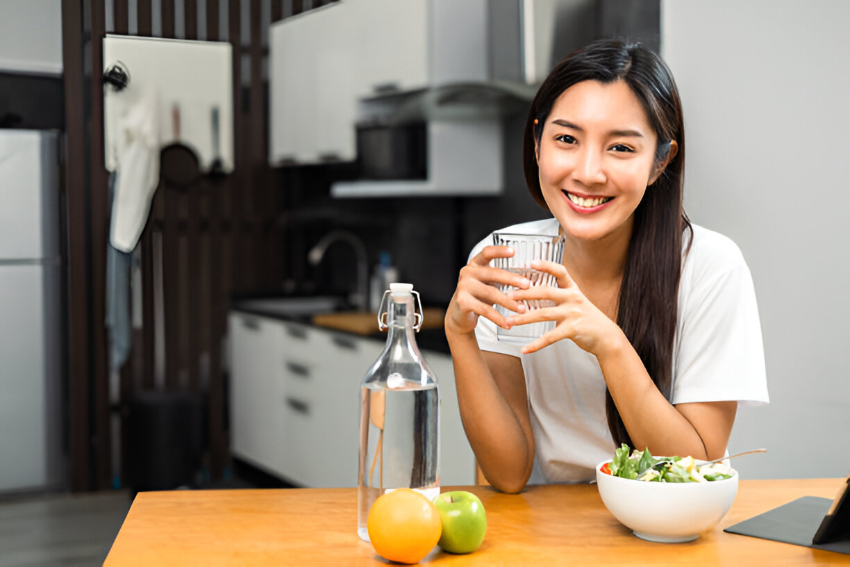 A woman at a table drinking water, accompanied by a bowl of colorful fruit